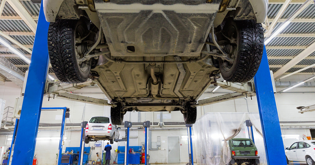 The underside of an SUV on a lift in a car mechanic's warehouse is visible. There are other cars in the background.